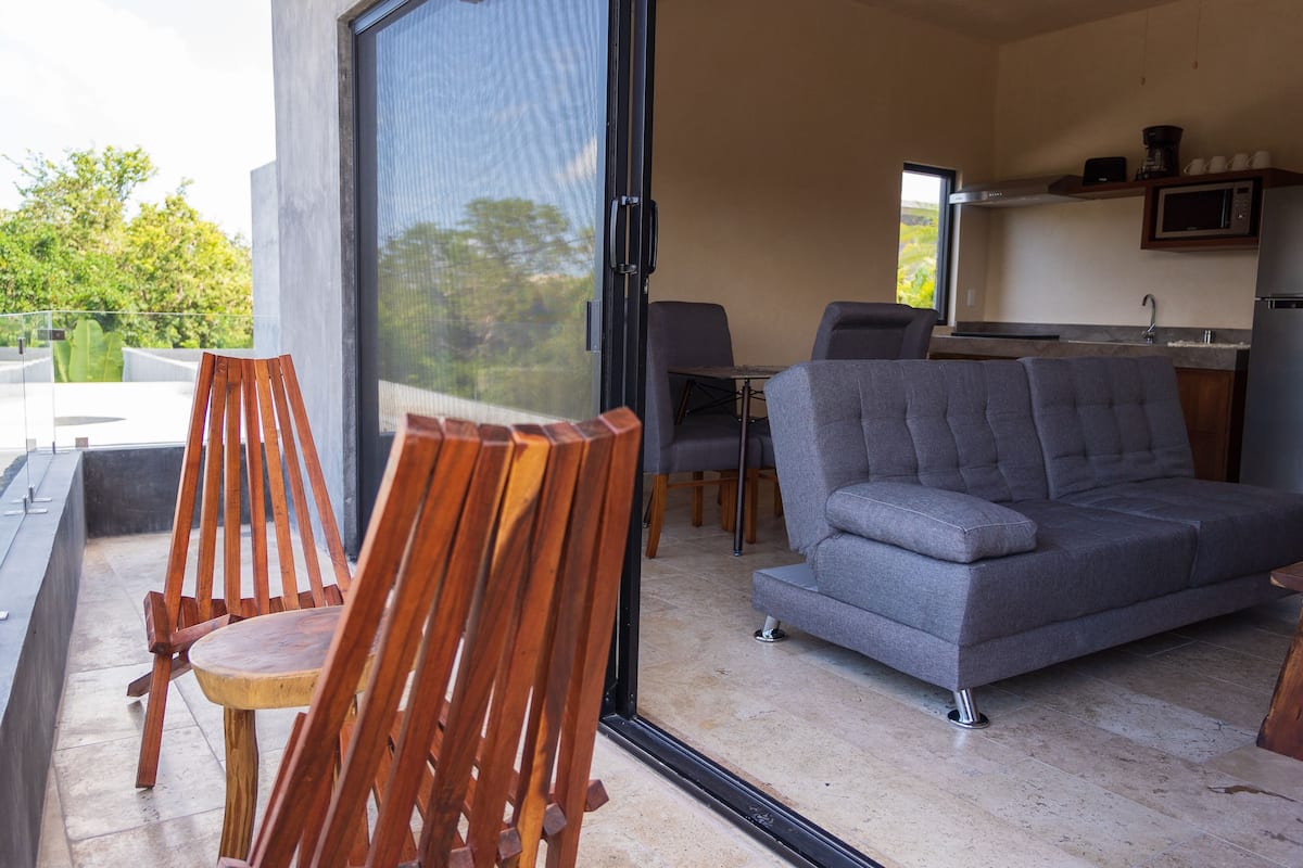 Una sala de estar moderna con un sofá gris, sillas de madera y un ventanal que da a un espacio exterior.