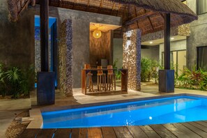 Un área de piscina moderna con bar, muros de piedra y plantas tropicales.
