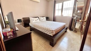 A bedroom with a bed, bedside tables, a desk, an air conditioner, a standing fan, and a window with curtains. - Ganesh House 2 Bedrooms (Tías)