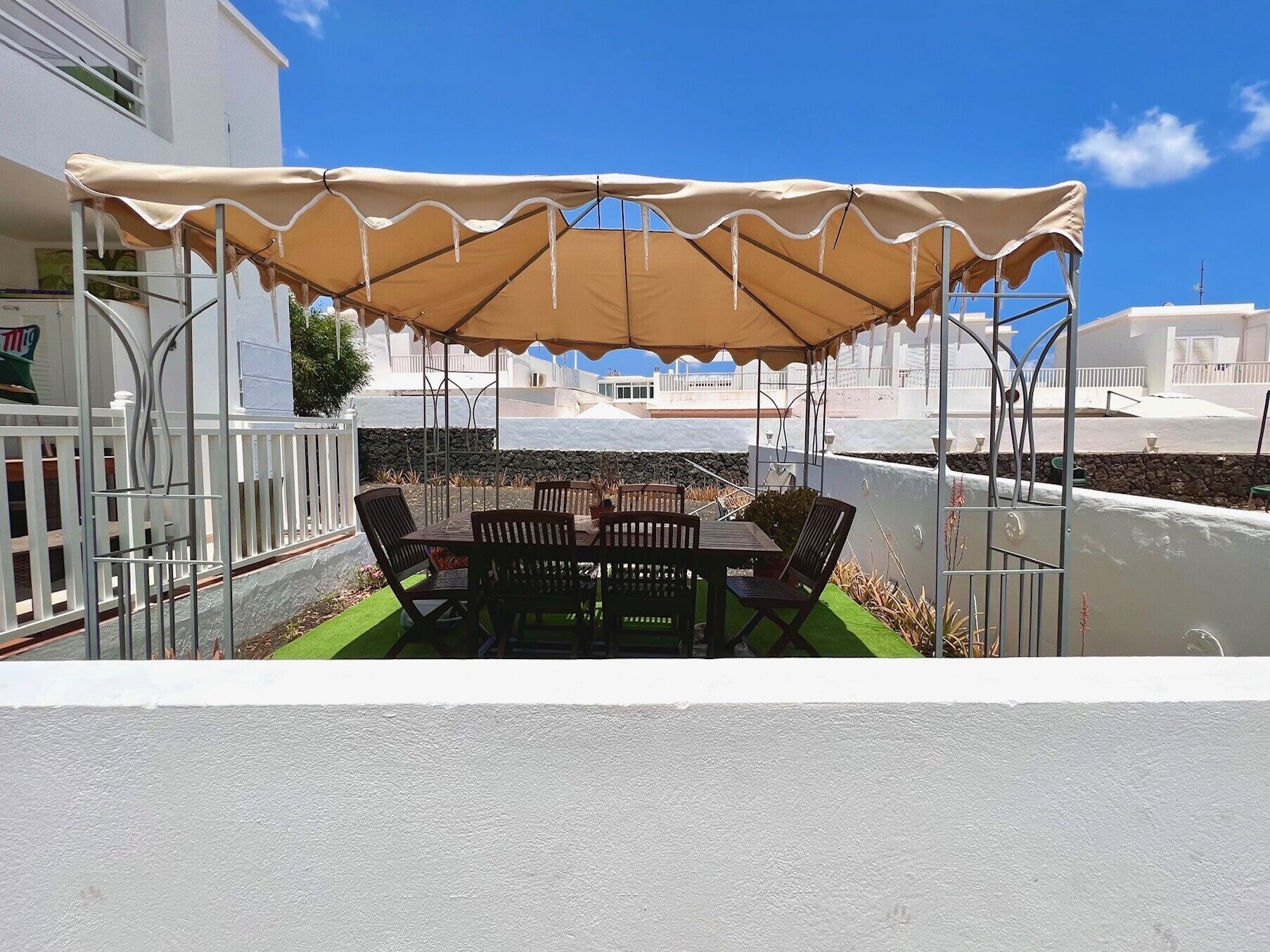 Une terrasse extérieure avec une table et des chaises, située sous une tonnelle et entourée d’une clôture blanche et d’un mur de pierres.