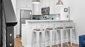 A modern kitchen with white cabinets, a stainless steel refrigerator, a microwave, and a bar area with white stools.