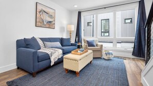 A living room with a blue sofa, a beige ottoman, a side table, and a lamp. There is a window with curtains and a painting on the wall.