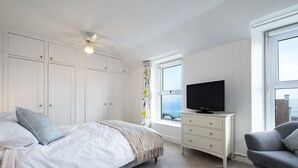 A bedroom with a bed, a TV on a dresser, a gray sofa, and a window with a view. - Seapinks (Port Isaac)