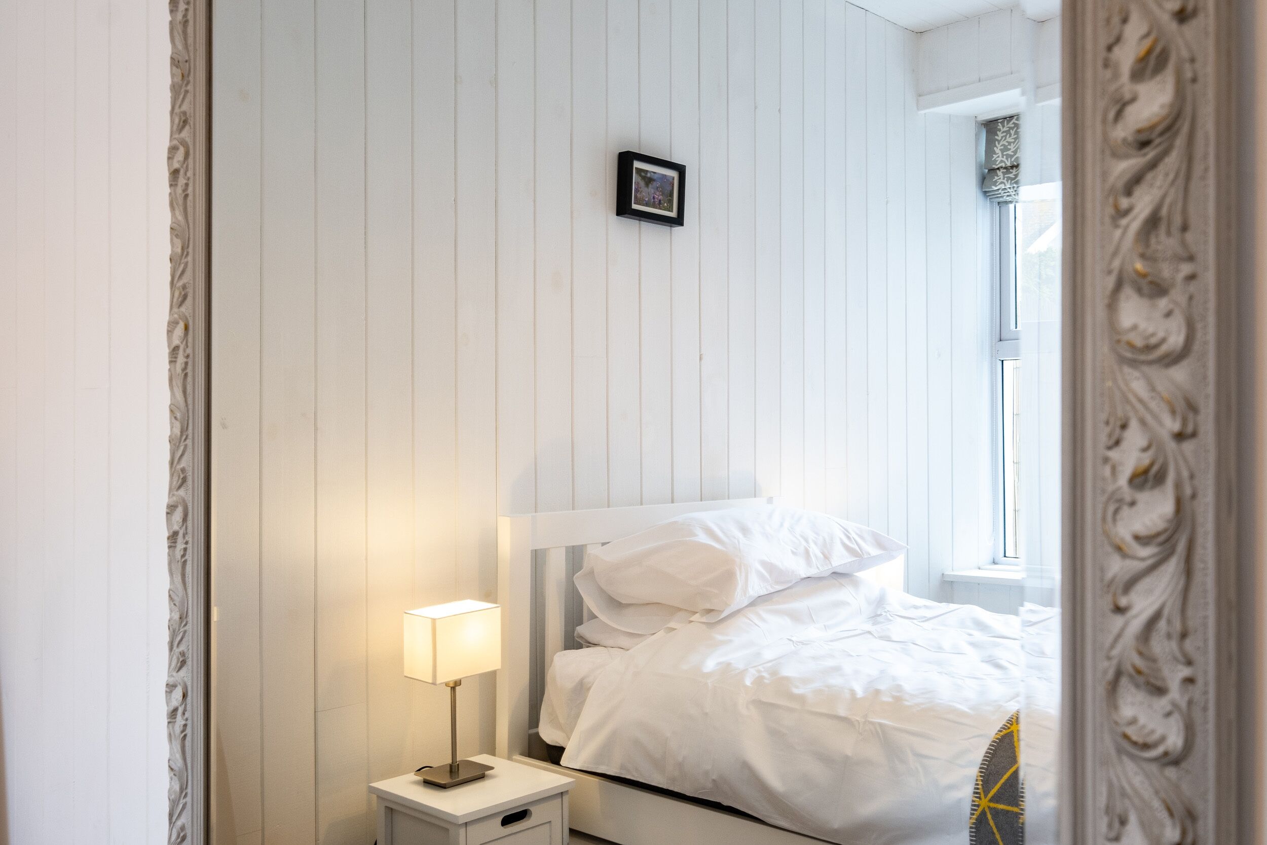 Een netjes opgemaakt bed met witte lakens, een nachtkastje met een lamp en een lijst met een foto aan de muur.