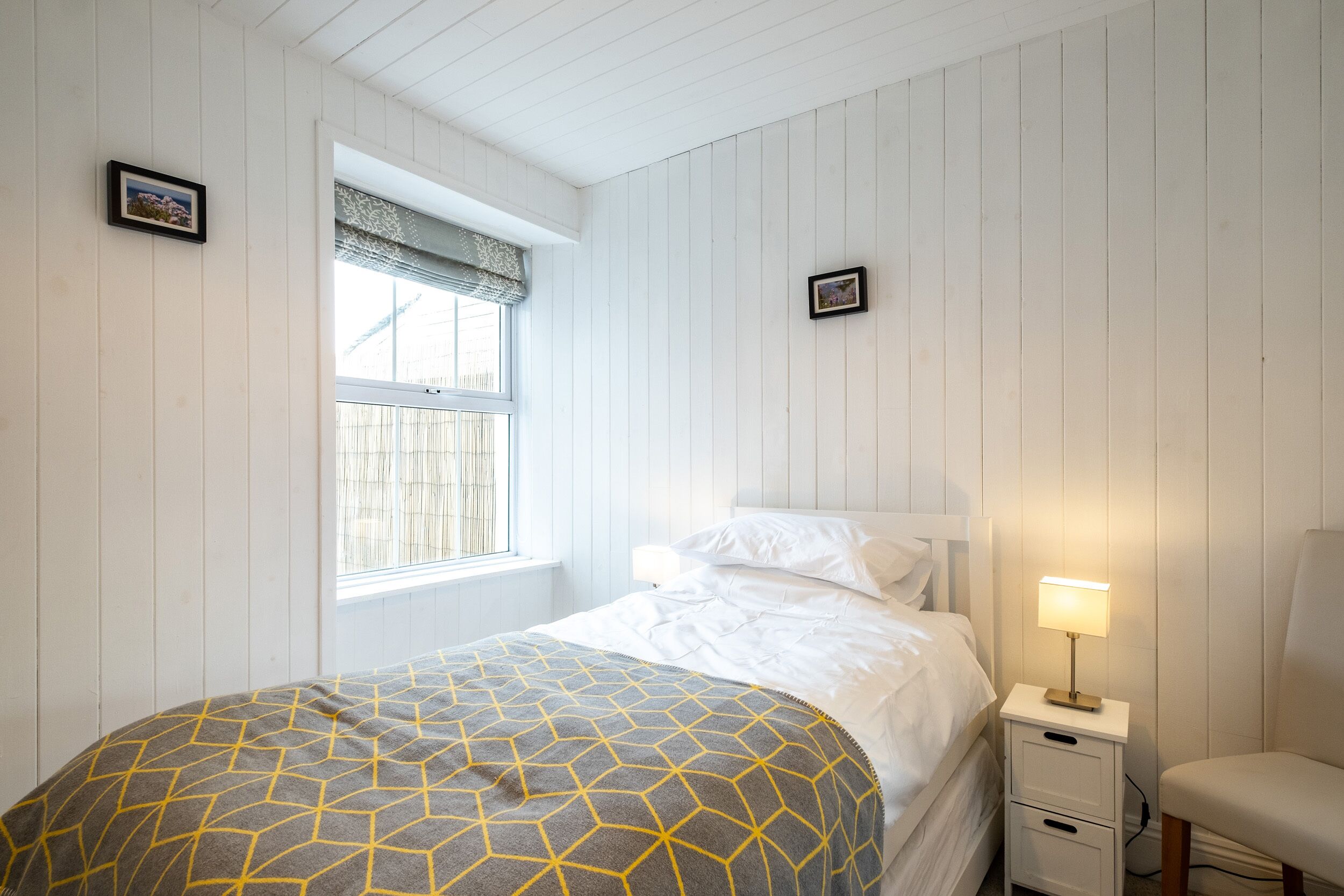 A bedroom with a bed, a nightstand with a lamp, a window with blinds, and a picture frame on the wall.