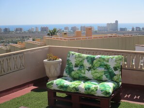 A balcony with a green and white patterned chair, a potted plant, and a view of the cityscape and ocean. - Apartamento Alemania Inmomart (Peñiscola)