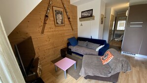 A cozy living room with a wooden sloped ceiling, a sofa, a small table, and a television. - Duplex N°5 Résidence Puy du Rocher (Le Lioran)