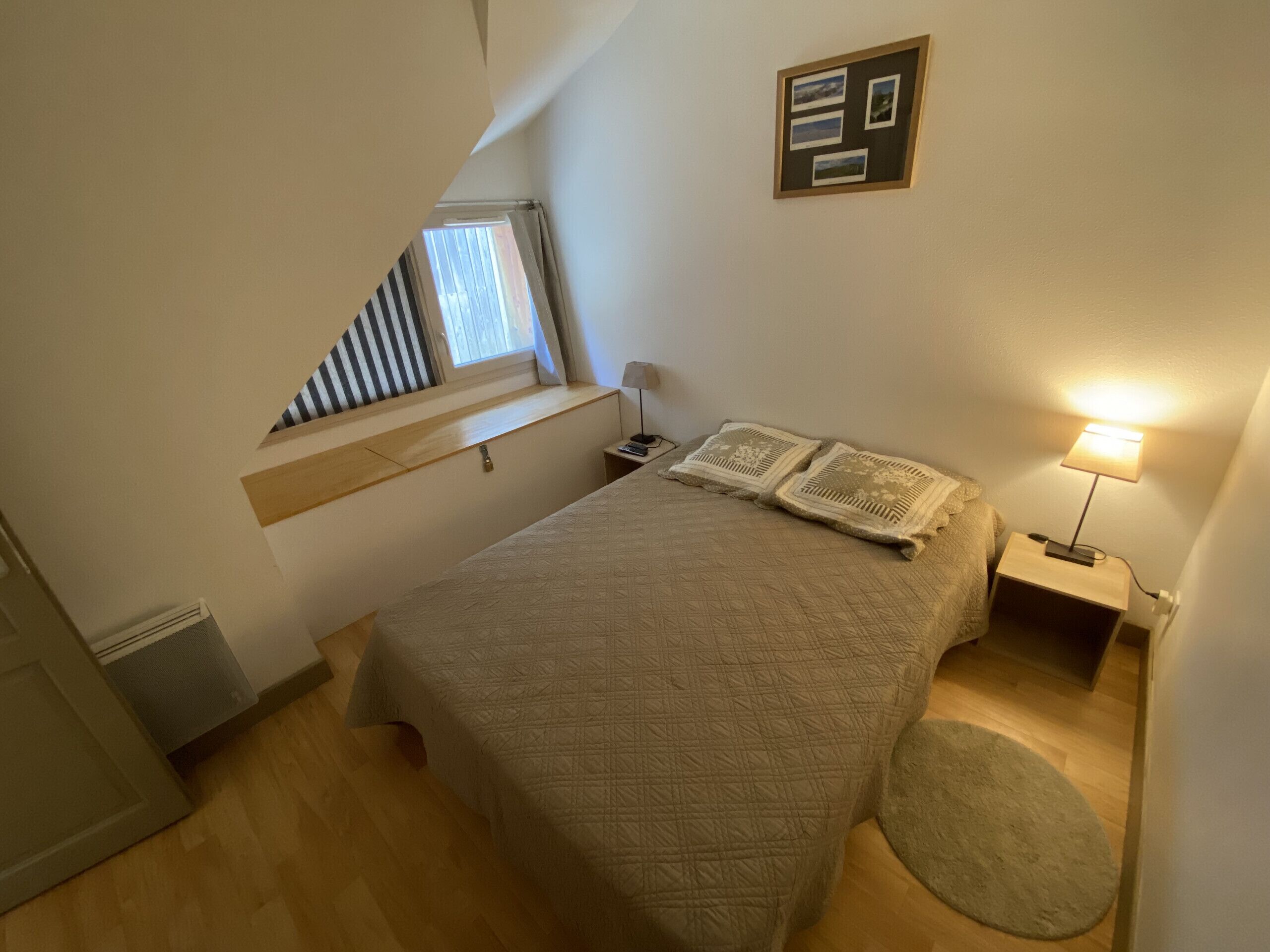 Een slaapkamer met een bed, een nachtkastje met een lamp, een raam met gordijnen en een aan de muur hangend fotolijstje.