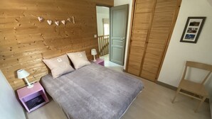 A bedroom with a wooden headboard, a bed with a grey blanket, two bedside tables with lamps, a wooden wardrobe, and a chair. - Duplex N°5 Résidence Puy du Rocher (Le Lioran)