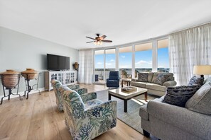 A spacious living room with a flat-screen TV, a ceiling fan, and a large window offering a city view.