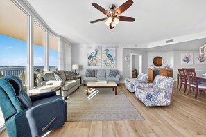 A spacious living room with a large window, a ceiling fan, and a dining area with wooden furniture. - PROFESSIONALY DECORATED, 3 BED 3 BATH UNIT!!! ASK ABOUT DISCOUNTS! (Orange Beach)