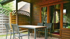 Une cabane en bois avec une table et des chaises sur une terrasse, un espace salon couvert et une porte donnant sur un jardin.