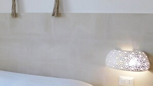 A neatly made bed with a beige bedspread, a white pillow, and a folded towel on the side. A wall-mounted lamp with a perforated shade is visible on the right side of the bed. - Monolocale Torre San Giovanni Verso Sud (Ugento)