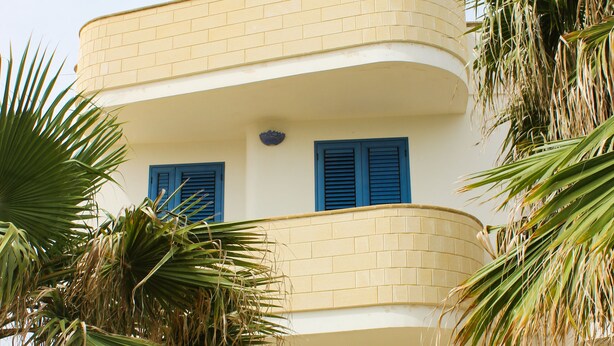 A two-story building with blue shutters, a curved balcony, and palm trees. - Monolocale Torre San Giovanni Verso Sud (Ugento)