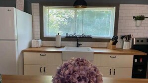 Een keuken met witte kastjes, een raam met zonwering, een spoelbak, een broodrooster en een potplant.