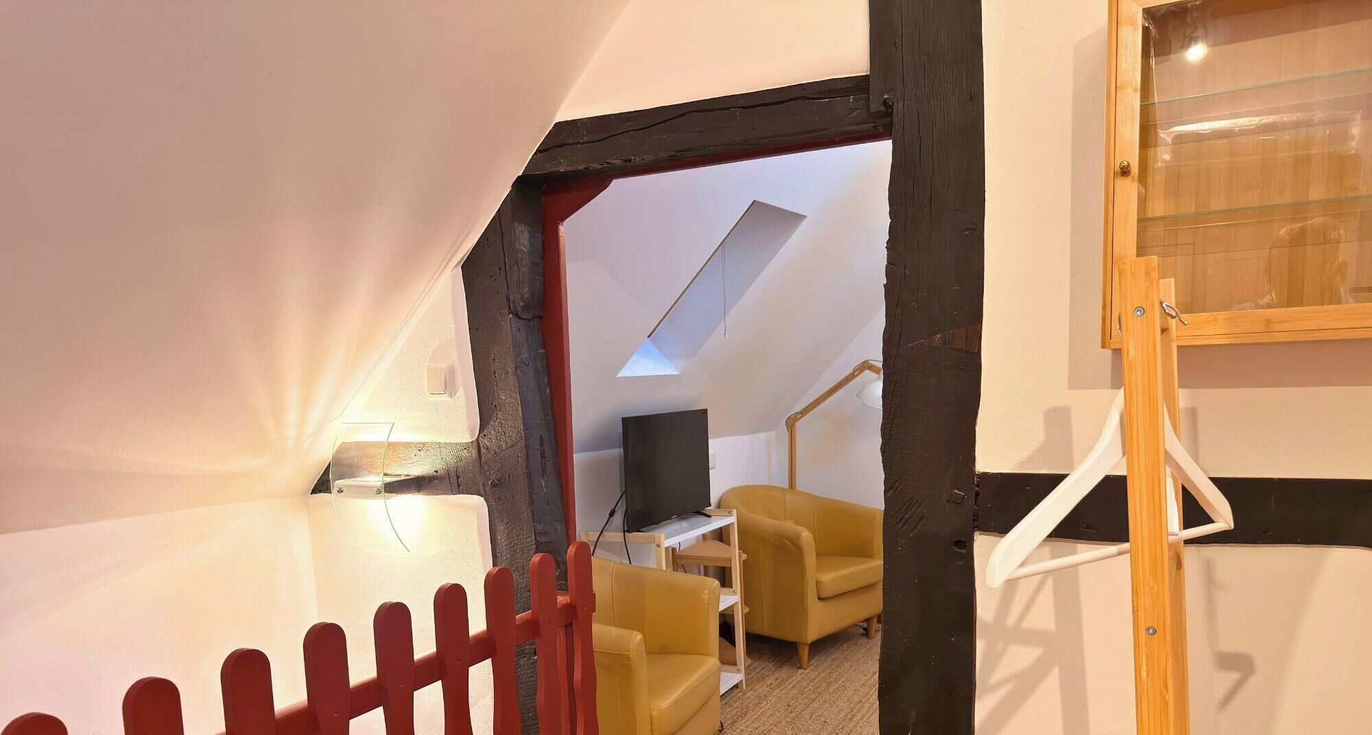 Quarto com teto inclinado, televisão em suporte, uma cadeira amarela e escada de madeira.