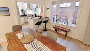 Un área de comedor con una mesa de madera, alfombra a rayas y dos sillas. Se ve una cocina al fondo con una ventana y un banco.