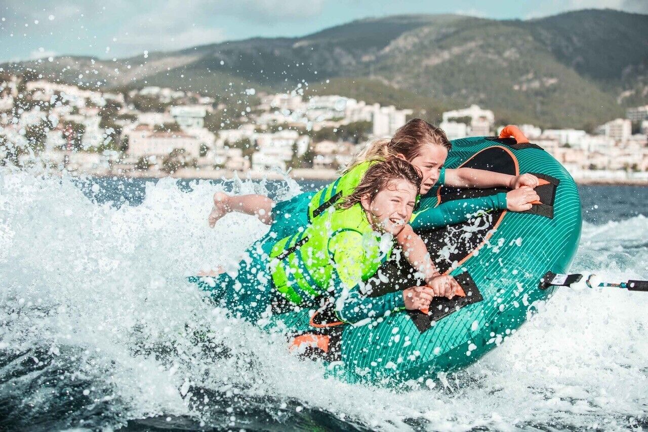 Two people in a green inflatable raft on water with a coastal town in the background.