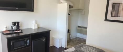 Habitación de hotel con televisor de pantalla plana en la pared, una kitchenette pequeña con microondas, cafetera y cajones de almacenamiento, un edredón estampado y un área de ducha visible a través de un umbral abierto.