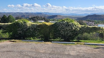 A gravel parking area with a hedge of white flowers and a view of distant hills and buildings.