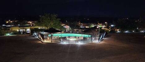 A fenced outdoor swimming pool area at night with surrounding buildings and streetlights.