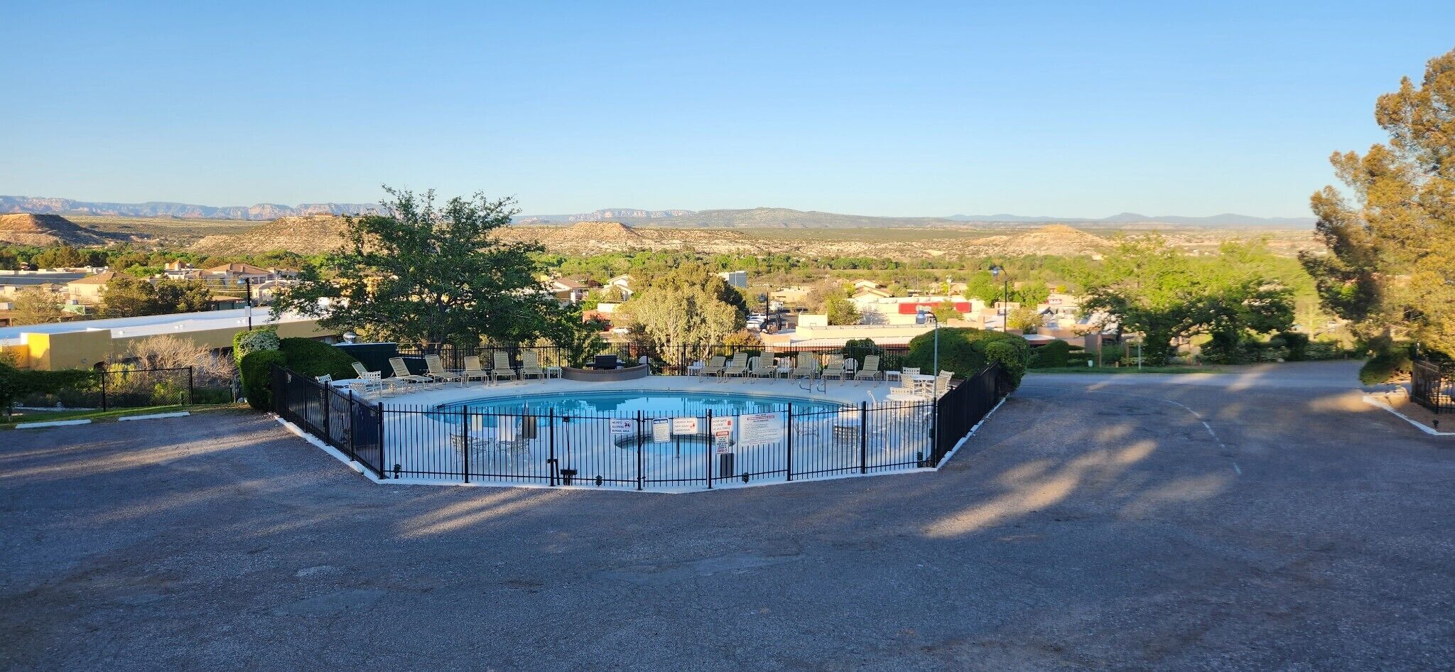 En swimmingpool omgiven av ett svart staket, med utsikt över ett bostadsområde och berg i bakgrunden.