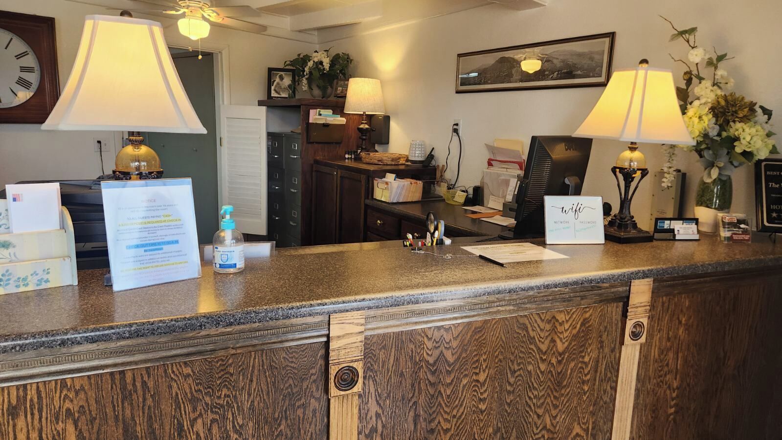 A hotel reception desk with a wooden counter, two lamps, a clock, a framed picture, and a vase with flowers.