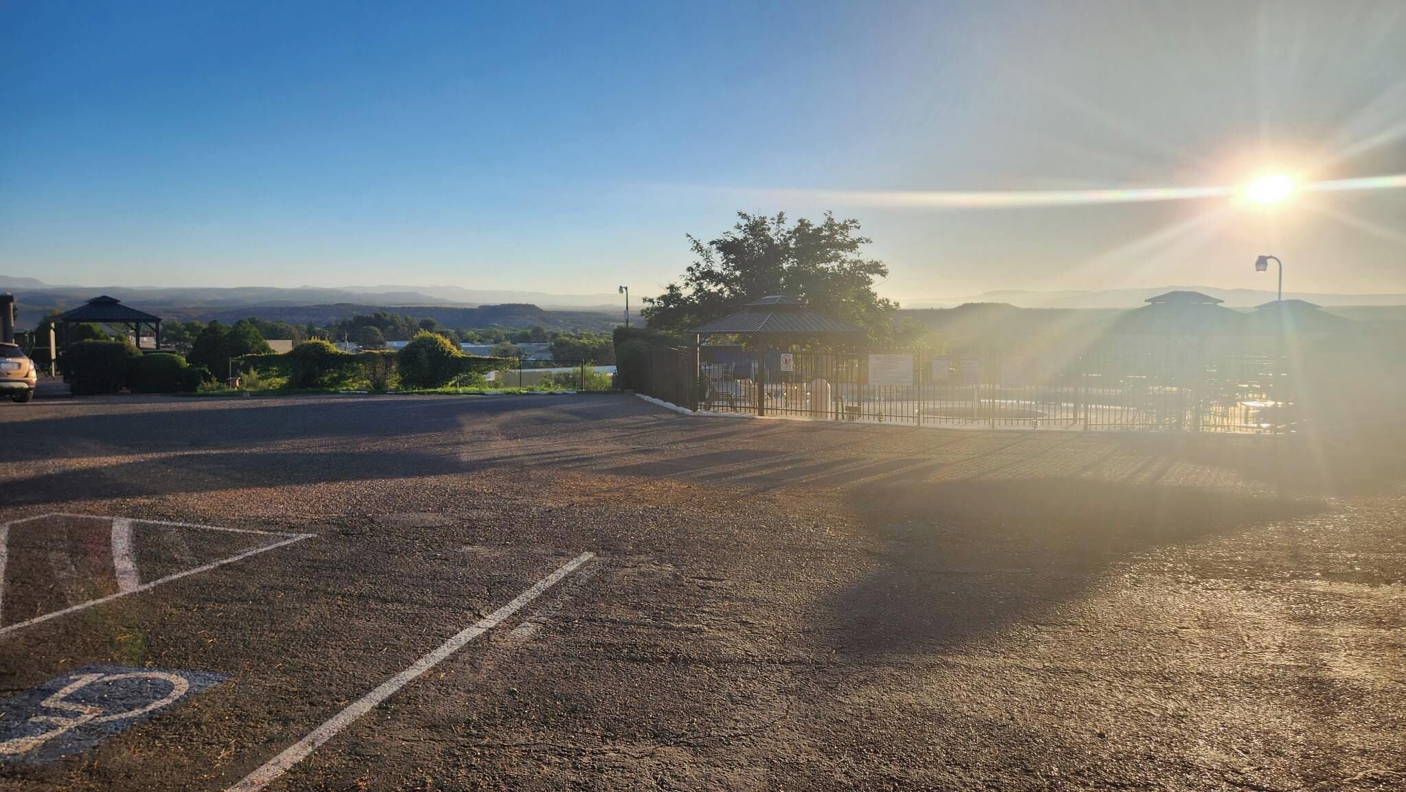 A parking lot with a gazebo and a fence, surrounded by greenery and distant hills.