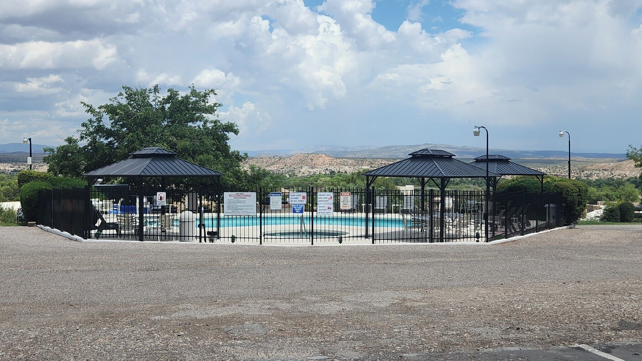 A fenced pool area with two gazebos, a clear sky, and distant mountains.