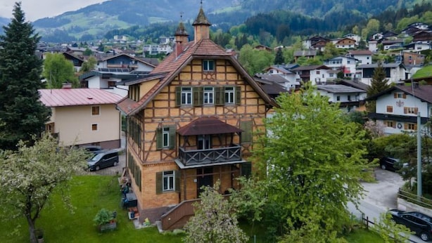 Une maison traditionnelle à colombages, dotée d’un balcon, entourée d’une végétation luxuriante et s’ouvrant sur un paysage montagneux.