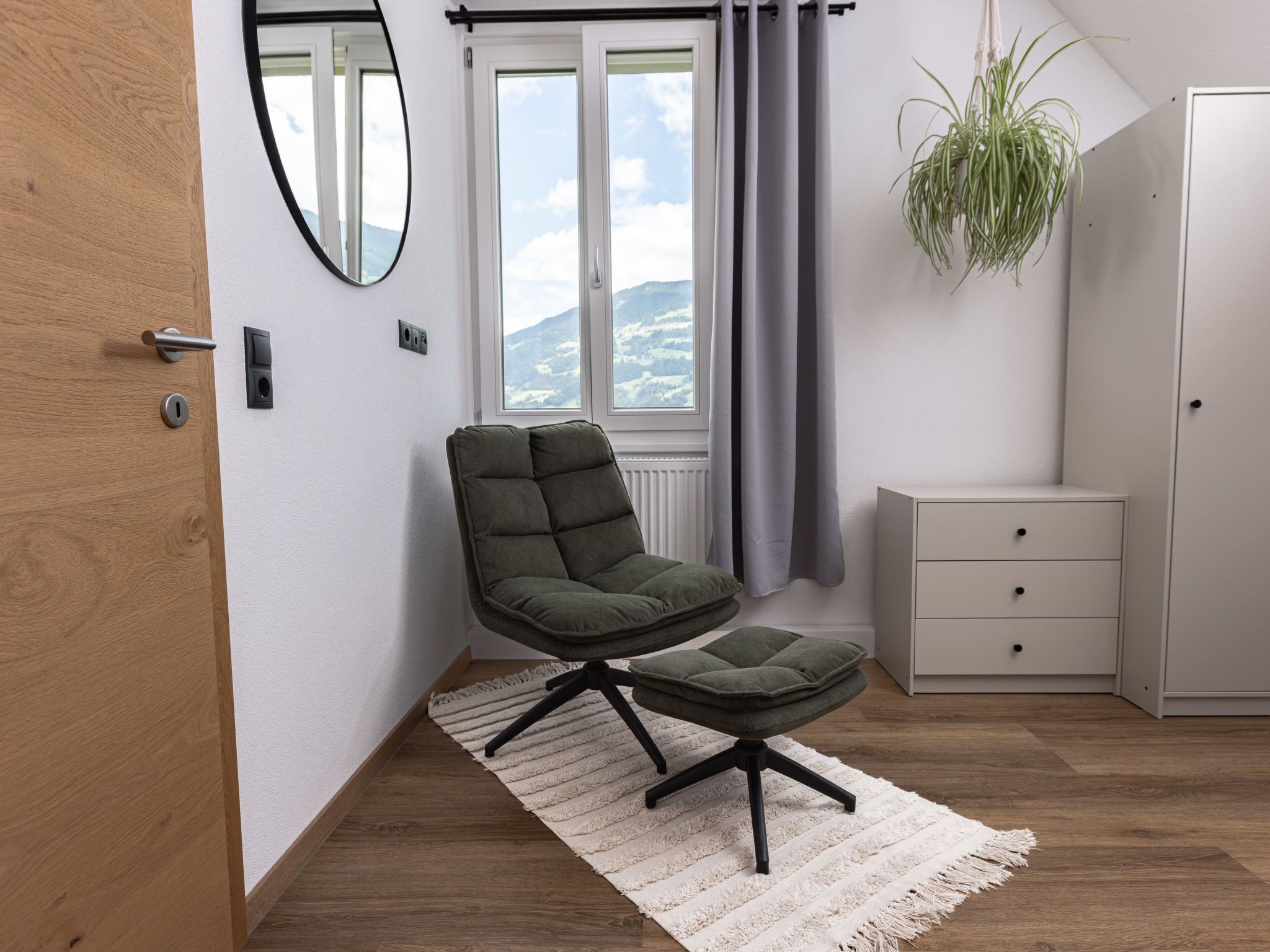 Ein modernes Zimmer mit einem grünen gepolsterten Sessel und Hocker, einem weißen Schminktisch, einer hängenden Pflanze und Bergblick durch ein Fenster.