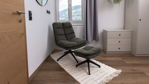 Une chambre moderne avec un fauteuil et un pouf verts, une commode blanche, une plante suspendue et une vue sur les montagnes par la fenêtre.