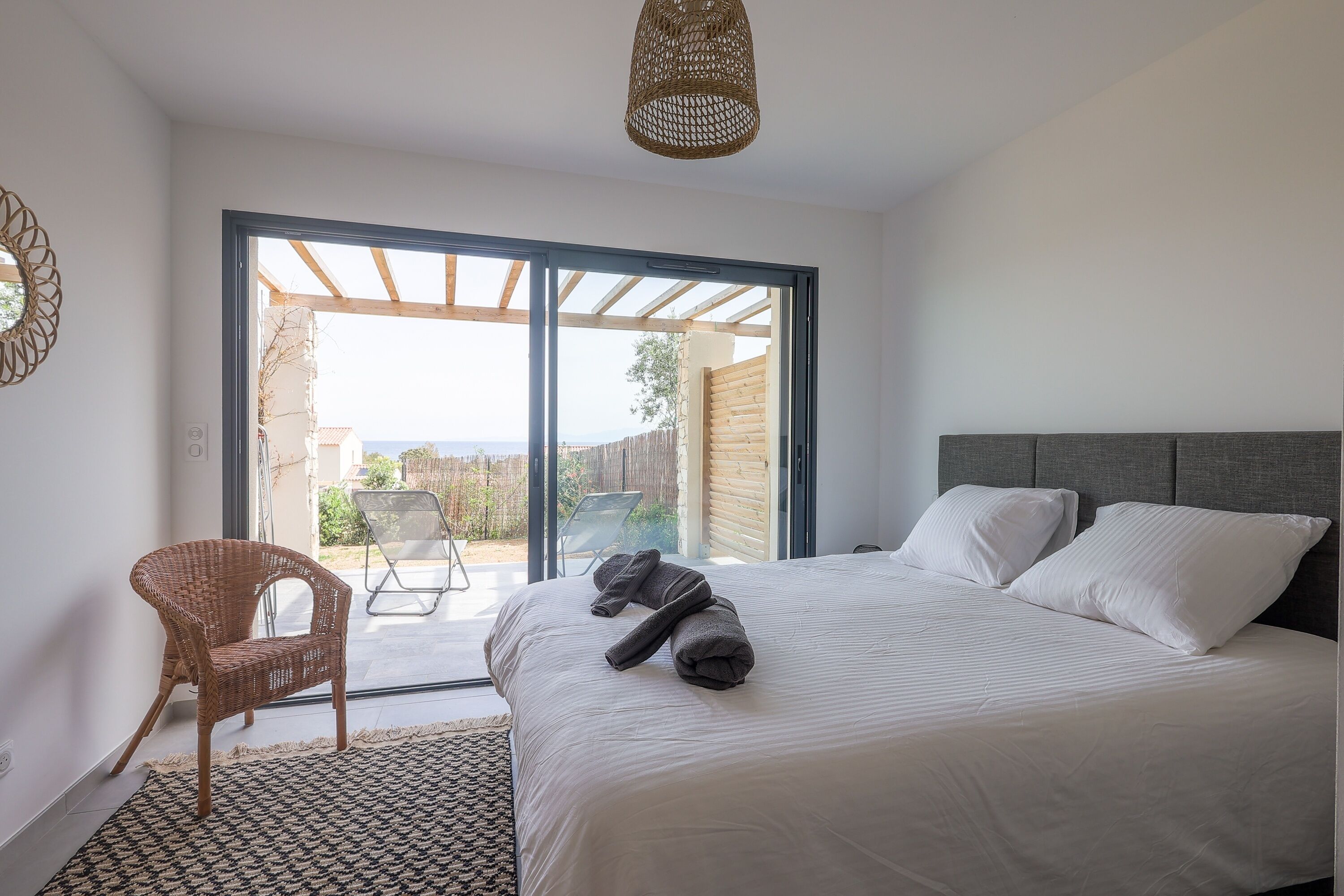 Un dormitorio con una cama grande, una silla de mimbre y vistas a un área exterior por una puerta corrediza de vidrio.