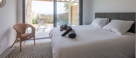 A bedroom with a large bed, a wicker chair, and a view through a sliding glass door to an outdoor area.
