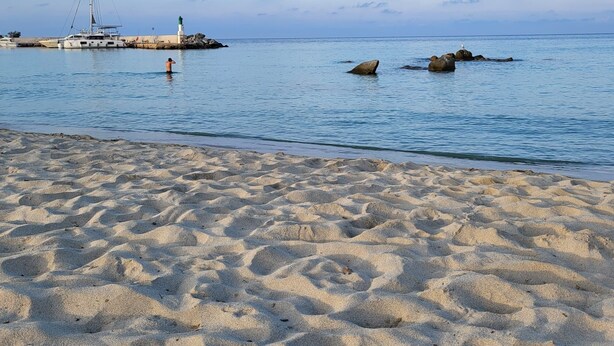 Una spiaggia sabbiosa con un mare calmo, un faro e una barca a vela all'orizzonte.