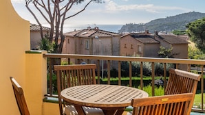 Un balcon avec une table ronde et deux chaises en bois, donnant sur un quartier résidentiel avec des maisons et offrant une vue sur la mer.