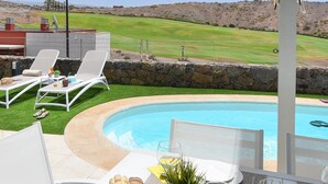 A poolside area with a table set for dining, lounge chairs, and a view of a golf course.