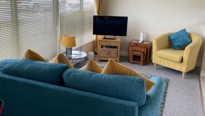 Een woonkamer met een blauwe bank, een gele fauteuil, een televisie en een kroonluchter.