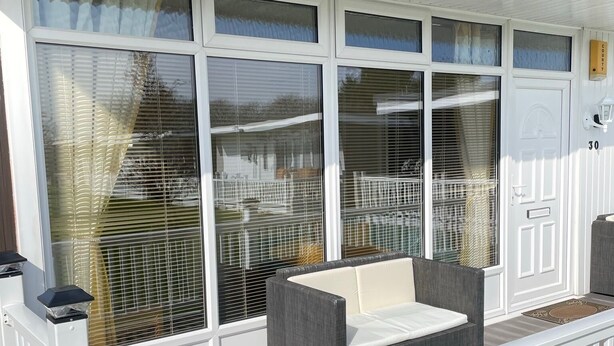Een overdekt terras met een witte balustrade, grote ramen met zonwering en een deur met een brievenbus.
