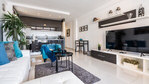 A modern living room with a white sofa, a coffee table, and a TV. There is a kitchen in the background with dark cabinets and a stove. - APARTMENT IN VIÑA DEL MAR - PLAYA DE LAS AMERICAS (Costa Adeje)