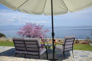Relax in style with a comfortable seating area under a spacious umbrella, overlooking a serene body of water and lush greenery.