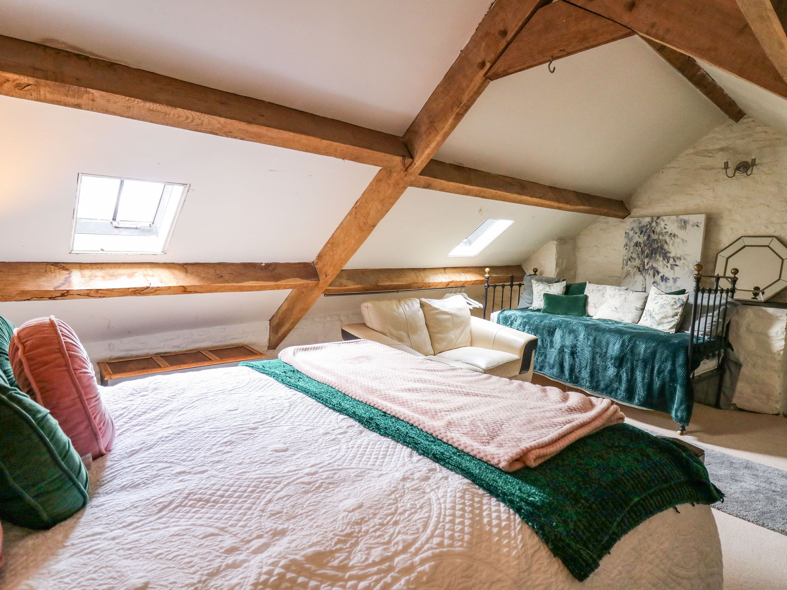 Une chambre mansardée chaleureuse avec un plafond en pente, des poutres en bois, un lit recouvert d’une couverture verte, une chaise et un tableau accroché au mur.