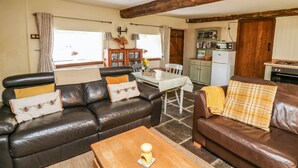 A cozy living room with a brown leather sofa, a wooden coffee table, and a dining area with a white table and chairs. A kitchen with a stove and cabinets is visible in the background. - The Bake House (Bridgend)