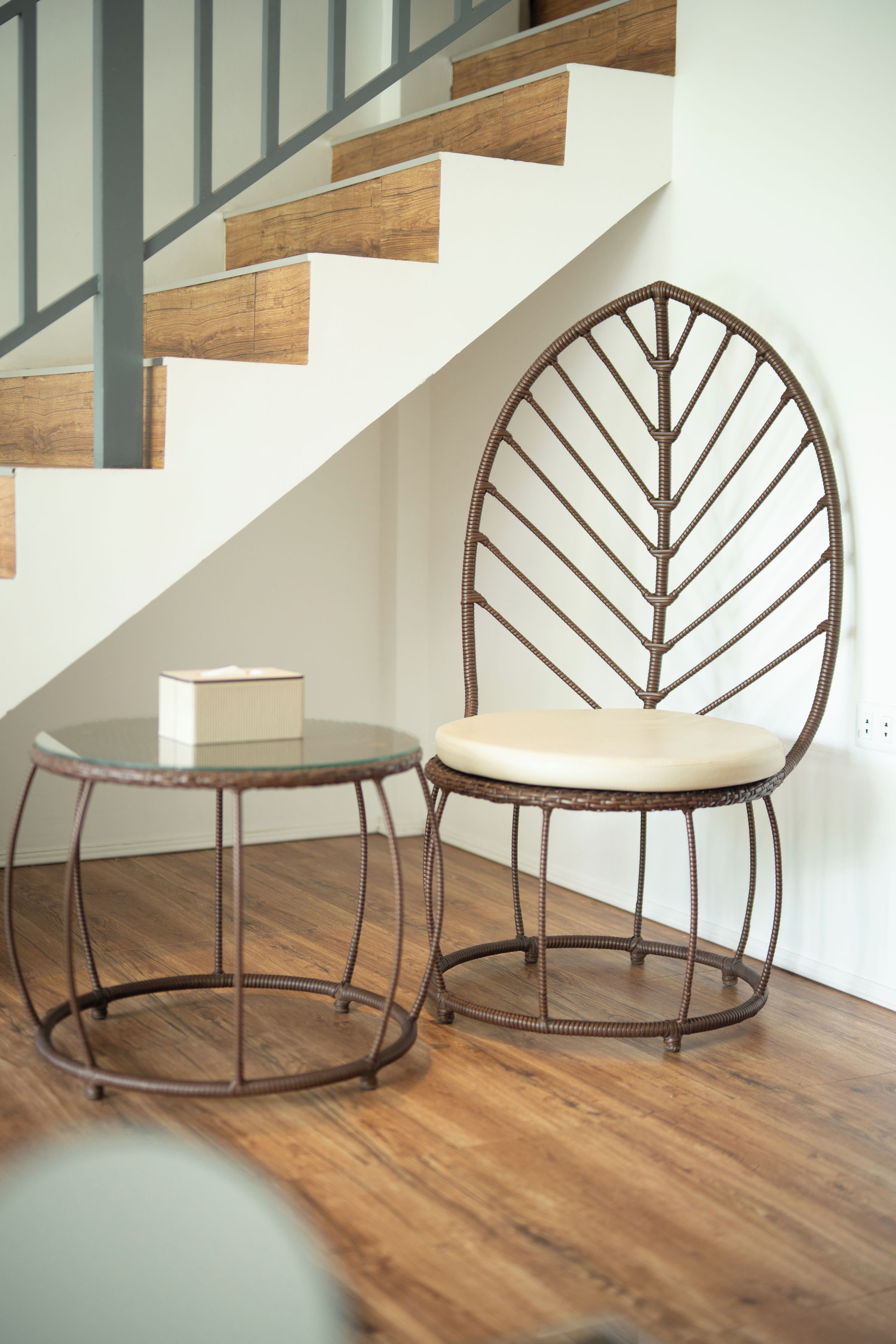 A chair with a leaf-like metal frame and a round table with a glass top, positioned near a staircase with wooden steps and metal railings.