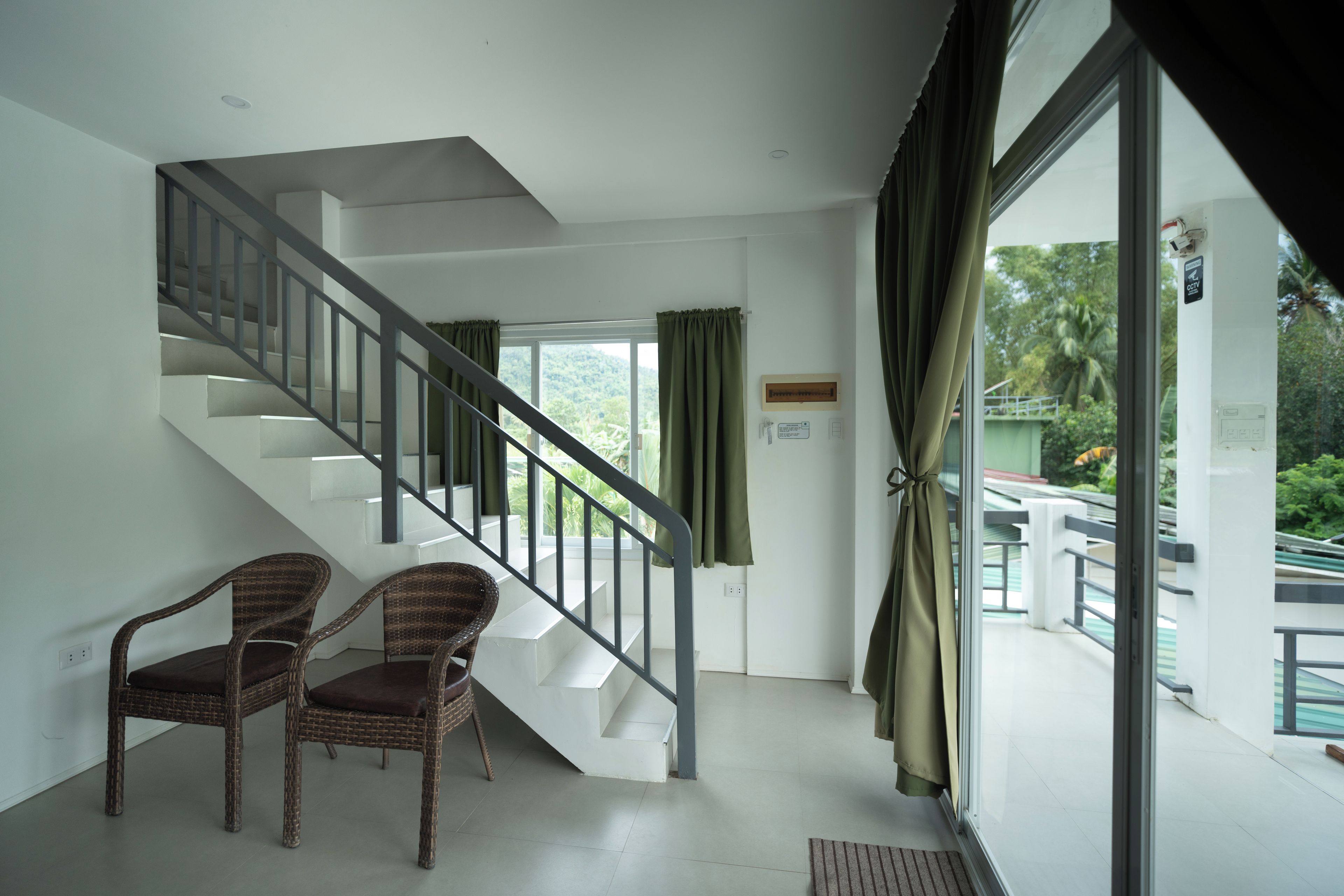 Una escalera con barandilla de metal que lleva a un piso superior, dos sillas de mimbre con cojines y vistas a zonas verdes a través de amplias ventanas.