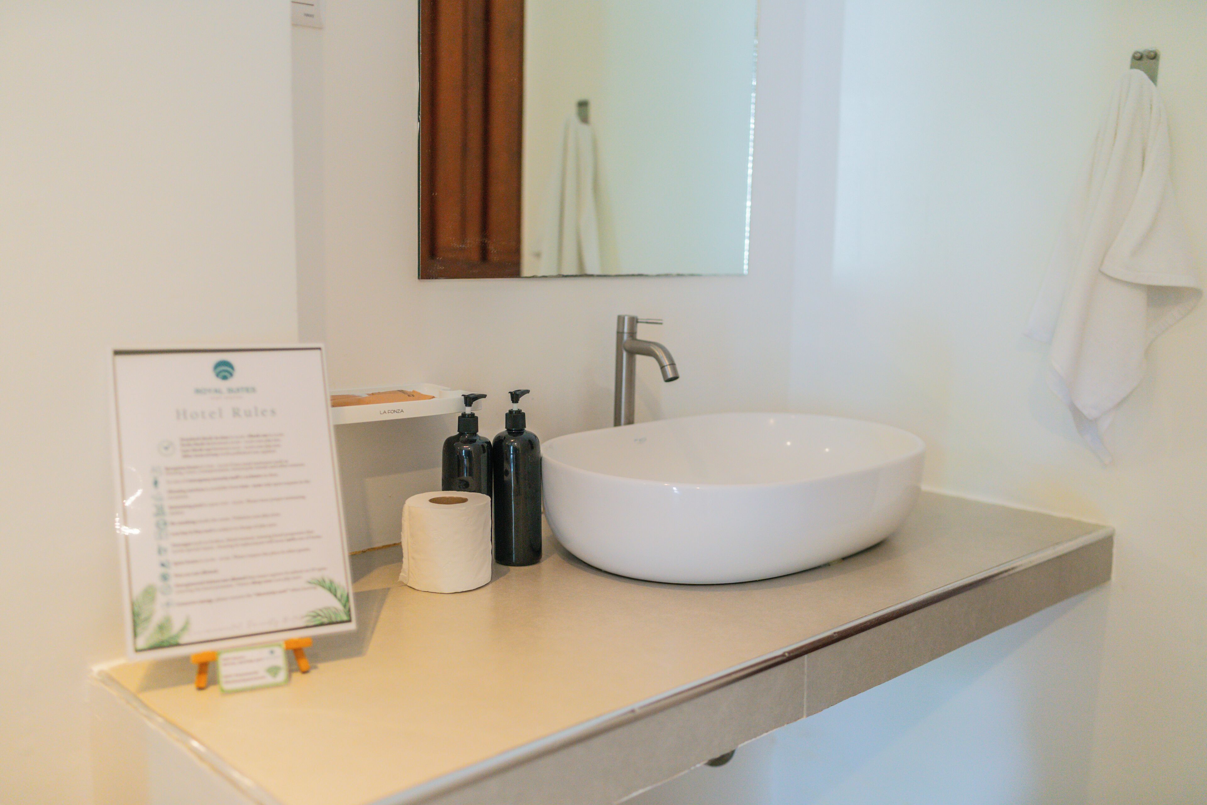 A bathroom with a large sink, a mirror, a towel rack, and a sign with hotel rules.