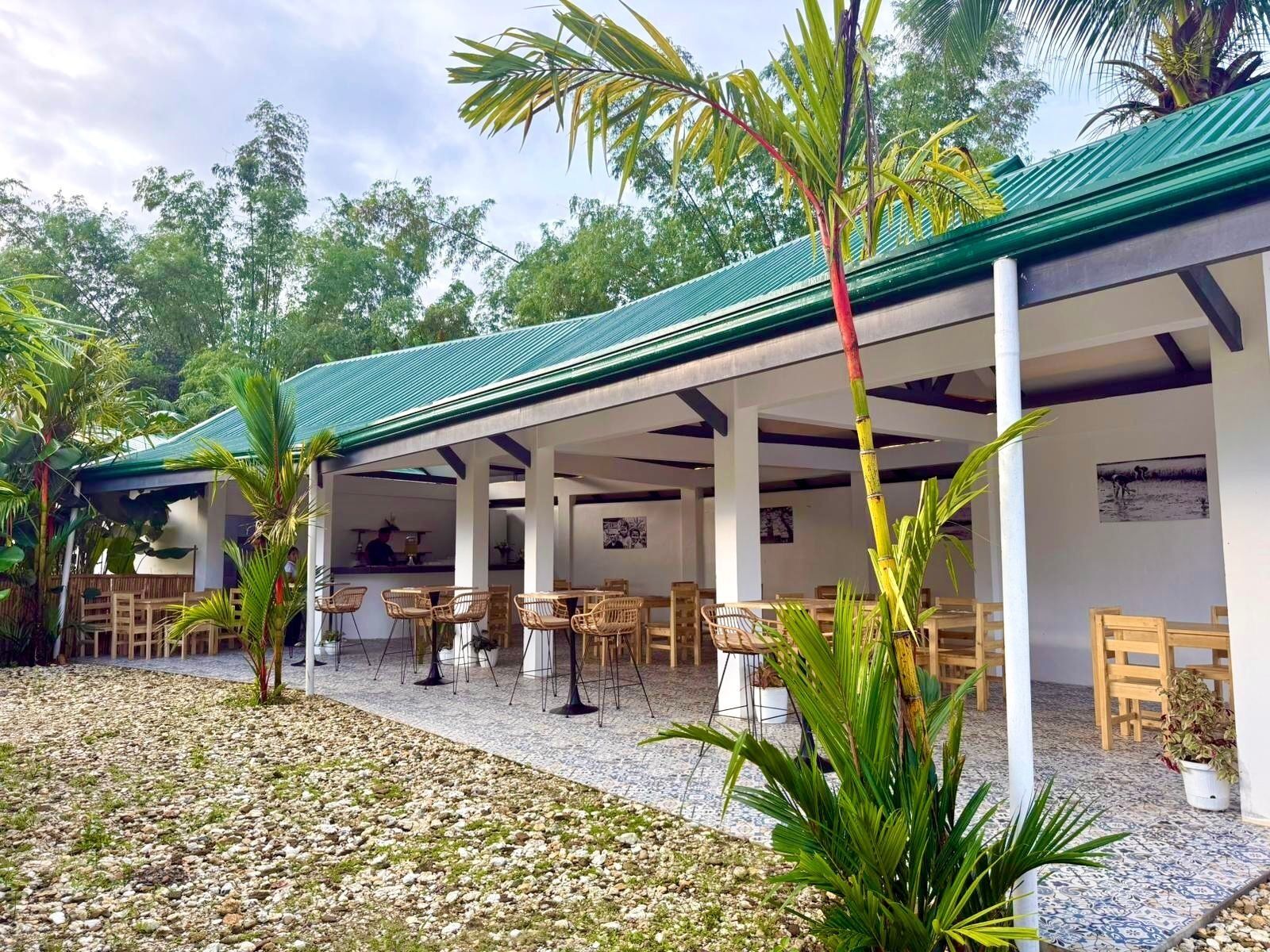 An outdoor covered patio with tables and chairs, surrounded by greenery and a tiled floor.