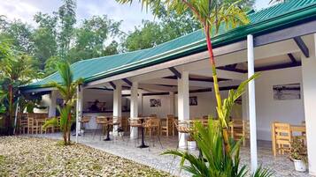 An outdoor covered patio with tables and chairs, surrounded by greenery and a tiled floor.