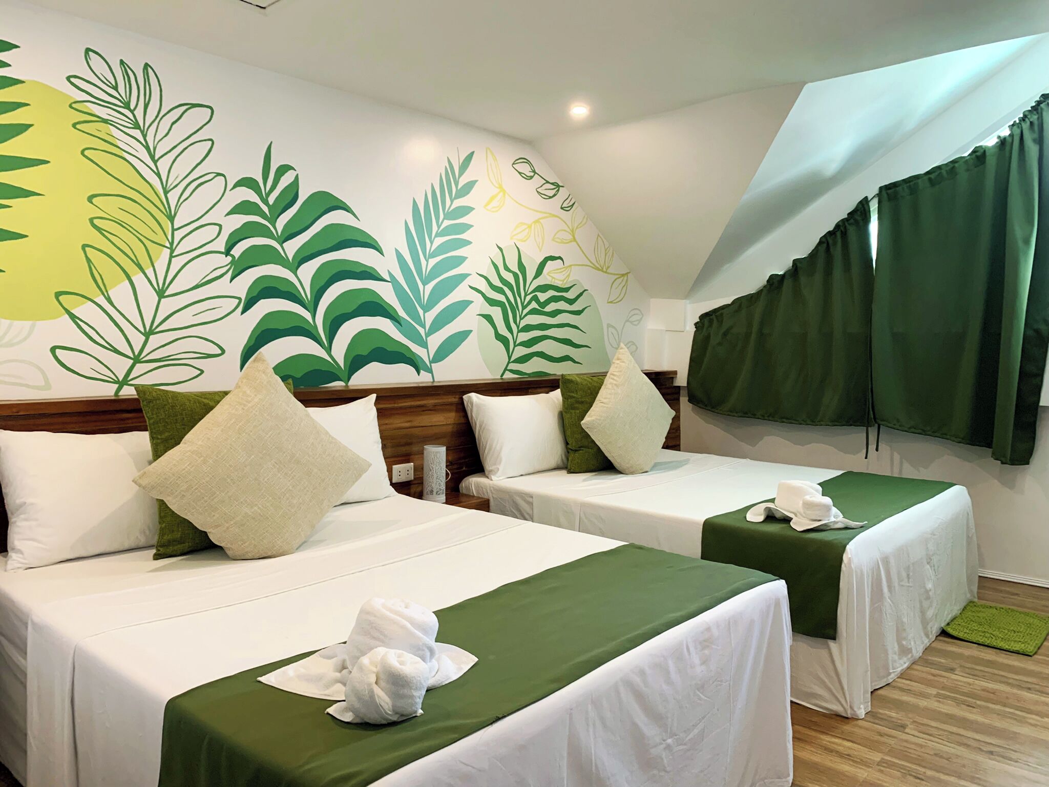 Habitación de hotel con dos camas, colchas verdes, almohadas blancas y un mural en la pared con motivos de hojas.