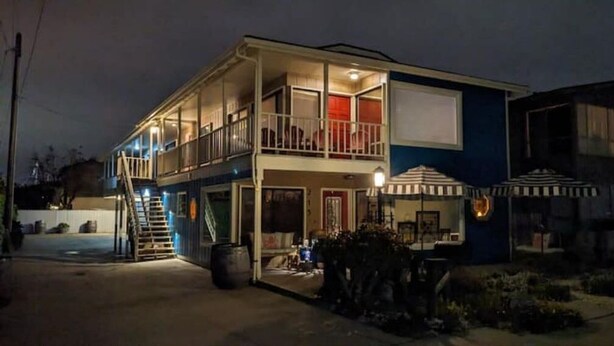 Un edificio de dos pisos con balcón, escalera y mobiliario exterior con sombrillas.